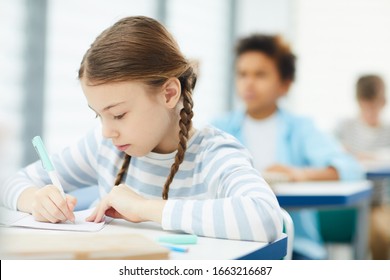 Horizontal medium close up portrait of Caucasian schoolgirl with two plaits sitting in modern classroom working on lesson task, copy space