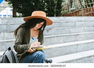 A horizontal medium full shot capturing a happy young adult woman sitting on outdoor concrete steps. Wearing a brown wide-brimmed hat, olive green cardigan, and blue jeans, she smiles while wearing wireless headphones, with her backpack and notebook resting beside her against a rustic urban wall.