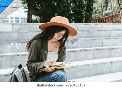 A horizontal medium full shot capturing a happy young adult woman sitting on outdoor concrete steps. Wearing a brown wide-brimmed hat, olive green cardigan, and blue jeans, she smiles while wearing wireless headphones, with her backpack and notebook resting beside her against a rustic urban wall.