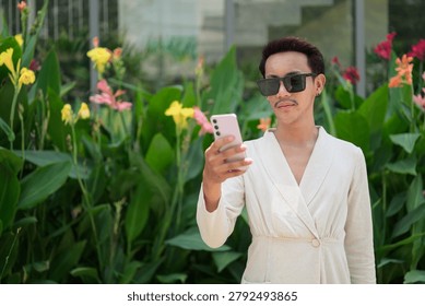 A horizontal medium portrait shot of a non-binary gay man of Isan, Thai descent standing outdoors. The model has short dark hair with vibrant reddish-maroon highlights, a mustache, black stud earrings on both ears, and is dressed in an off-white or cream-colored wrap blazer jacket with a deep v-neck line. Positioned on the right side of the frame, the individual is wearing large square black sunglasses and is looking down with a neutral, focused expression while holding and viewing a lavender-pink smartphone in their right hand. Immediately behind the subject on the left is a vibrant bed of large-leaf green tropical canna lily plants featuring colorful yellow and pink blossoms. In the background, a modern building facade with large glass windows reflects the ambient outdoor daylight. Shot with natural, soft diffused sunlight highlighting the model's authentic, unretouched skin texture and the woven linen-like pattern of the garment. The composition features an off-center placement with a very shallow depth of field, keeping the individual sharply in focus while smoothly blurring the botanical background on the left side of the frame.
