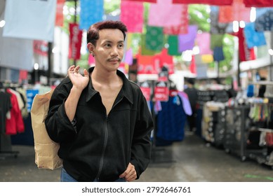 A horizontal medium portrait shot of a non-binary gay man of Isan, Thai descent standing outdoors in a bustling street market aisle. The model has short dark hair with subtle reddish highlights, a mustache, black stud earrings, and is dressed in a casual black jacket. Positioned on the left side of the frame, the individual is looking away off-camera to their right with a calm, neutral expression while carrying a brown paper shopping bag held over their shoulder. The background features blurred clothing racks, market stalls, and colorful banners hanging overhead, all softly out of focus. Shot with soft, natural outdoor daylight that defines the model's authentic, unretouched features. The composition places the subject off-center with a shallow depth of field, keeping the individual sharp while smoothly blurring the background elements.