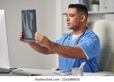 Horizontal Medium Portrait Shot Of Serious Middle-Eastern Male General Practitioner Sitting At Desk In Office Looking At X-ray Shot