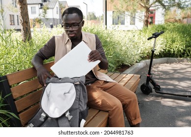 Horizontal Medium Portrait Of Young Black Man Wearing Casual Clothes With Eyeglasses Spending Summer Morning Outdoors Sitting On Bench Taking Laptop Out Of Backpack