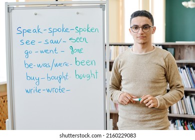 Horizontal medium portrait of young Middle-Eastern student standing at whiteboard with irregular verbs written on it looking at camera