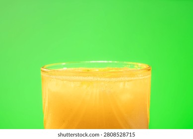 Horizontal medium shot centered on the upper portion of a textured glass filled with yellow juice, ice, and bubbles, located beneath a wide solid green background.