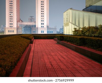 Horizontal night view of an empty red wooden paved pathway bordered by manicured hedges, directing attention to massive glowing twin commercial high-rise buildings. Illustrates urban scale, property development, and corporate architecture.