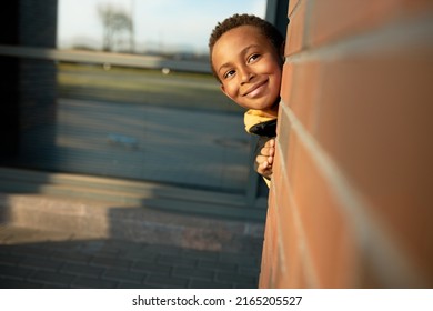 Horizontal outdoor image of happy funny african american kid playing hide and seek in city streets, hiding behind red brick wall from school classes, smiling. Carefree childhood. Happy kids