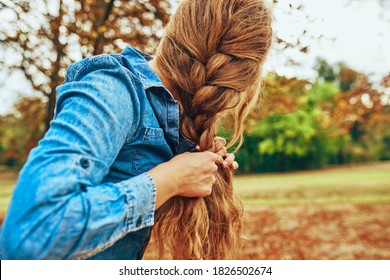 Horizontal outdoor image of a young woman arrange her red hair, making the braid, wearing a blue denim shirt, posing on nature background.  - Powered by Shutterstock