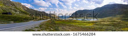 Horizontal panoramic view of winding road along mountains and lake. Breathtaking mountain landscape of the Norwegian nature. Ultimate road trip destination to Scandinavia. Green moss clouds blue sky. 