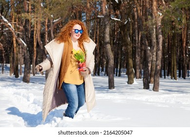 Horizontal photo of amused curly haired woman wearing warm clothes and blue sunglasses while keeping yellow snowdrops and walking through snowdrifts in sparse forest