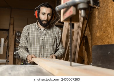 Horizontal photo an attentive carpenter man mid adult caucasian with protective ear muffs guides a wooden board through a bandsaw, demonstrating precision in his craft. Business concept. - Powered by Shutterstock