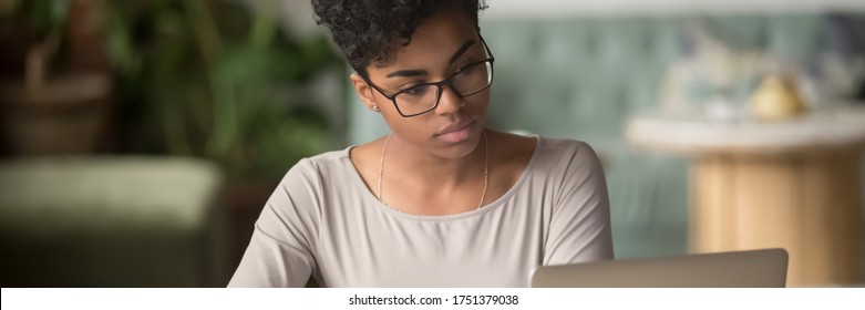 Horizontal photo banner for website header design, African student girl wear glasses sit indoors look at pc screen study on-line, do assignment, prepare for university admission exams, e-study concept - Shutterstock ID 1751379038