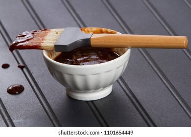 Horizontal Photo Of Barbecue Sauce With Wooden Brush And White Cup Dripping On A Black Table In A White Dish On A Black Table Top With Diagonal Lines