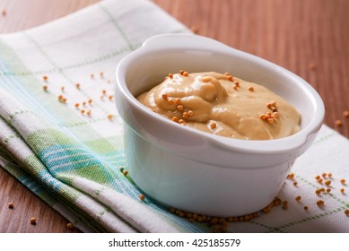 Horizontal photo of bowl with mustard. White oval bowel full of orange mustard. Mustard seeds spilled in and around bowl. Oval bowl on green checkered towel. Towel on wooden board. - Shutterstock ID 425185579