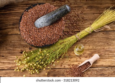 Horizontal photo with a bunch of flax plants with dried blooms bonded by natural cord placed on old wooden board and with burlap around which is near to bottle with flax oil and heap of flax seeds.