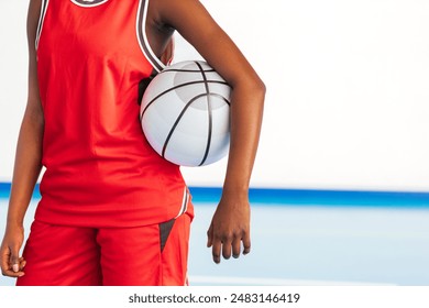 Horizontal photo close-up of a basketball player in a red uniform holding a basketball under their arm on an indoor court, emphasizing focus, determination, and sportsmanship in a competitive setting. - Powered by Shutterstock