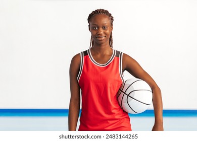 Horizontal photo a confident young female basketball player in a red uniform holds a basketball under her arm on an indoor court, exuding determination and readiness for the game ahead. Sport concept. - Powered by Shutterstock