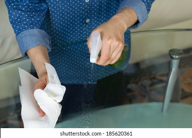 Horizontal Photo Female Hands Spraying Cleaning Solution, From Spray Bottle, Onto Dirty Glass Table With Paper Towels And Sofa In Background 