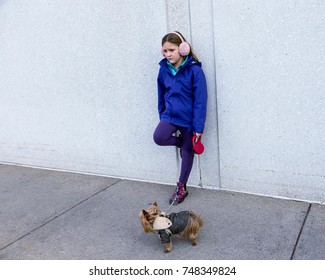 Horizontal Photo Of Forlorn-looking Little Girl In Fall Jacket And Ear Muffs Leaning With One Foot On Wall While Holding Tiny Yorkshire Terrier Dog On Leash