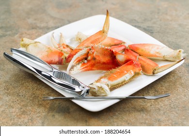 Horizontal Photo Of Freshly Cooked Dungeness Crab Legs, Focus On Large Claw In Center, On White Plate With Stainless Crab Crackers And Stone Counter Top Underneath  