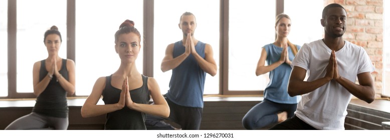 Horizontal photo group young multiracial people girls guys practicing yoga standing in Vrksasana Tree pose hands in namaste gesture. Healthy lifestyle wellness concept banner for website header design - Powered by Shutterstock