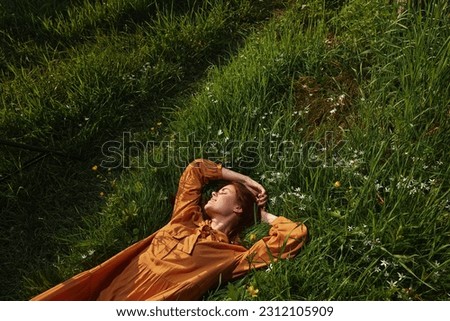 horizontal photo of a happy, relaxed woman, resting lying in the grass, in a long orange dress, with closed eyes and a pleasant smile on her face, enjoying harmony with nature and recuperating