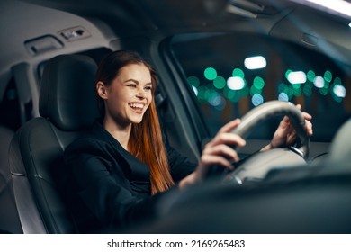 Horizontal Photo Of A Nice Woman In A Black Shirt Driving A Car While Sitting At The Wheel With A Pleasant Smile On Her Face At Night