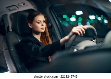 Horizontal Photo Of A Nice Woman In A Black Shirt Driving A Car While Sitting At The Wheel With A Pleasant Smile On Her Face At Night