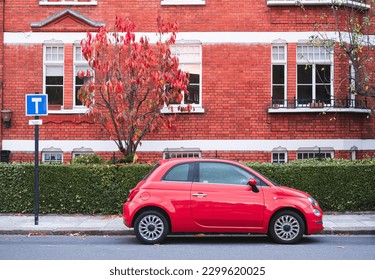 Horizontal photo of a red small car parked in front of a red brick building with a red tree on the streets of London, United Kingdom. - Shutterstock ID 2299620025