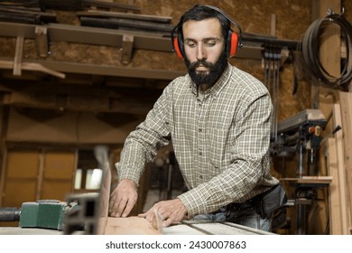 Horizontal photo serious carpenter with hearing protection operates a table saw, pushing a wooden board towards the rotating blade in a busy woodshop. Business concept. - Powered by Shutterstock