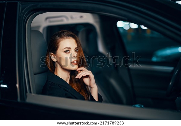 horizontal photo\
from the side, at night, of a woman sitting in a black car and\
thoughtfully looking at the camera holding her hand near her face,\
parking the car in the parking\
lot