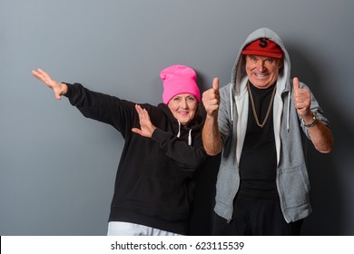 Horizontal photo of two old people dancing in front of a gray wall. Grandad shows thumbs up, granny raised her hands aside. Funny grandparents in modern clothes have a good time.