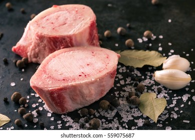Horizontal photo with two pieces or slices of big marrowbone with few drops of blood and meat on side. Bones are placed on worn black baking tray with salt and other spices around.