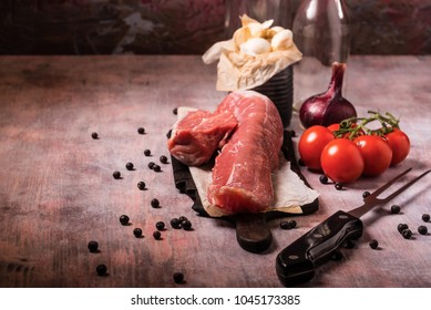 Horizontal Photo Of Whole Pork Tenderloin. Raw Meat Is On Vintage Wooden Board With Big Fork, White Garlic In Old Can And Few Red Cherry Tomatoes. Two Empty Bottles Are In Background.