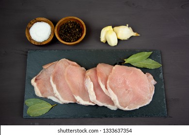 Horizontal Photo Of Whole Pork Tenderloin Raw Meat Is On A Black Slate Dish With Pepper Salt Aromatic Herbs And White Garlic