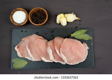 Horizontal Photo Of Whole Pork Tenderloin Raw Meat Is On A Black Slate Dish With Pepper Salt Aromatic Herbs And White Garlic