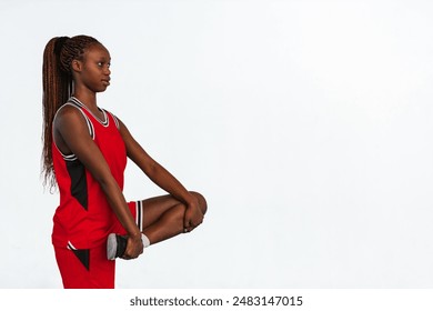 Horizontal photo A young female athlete in a red sports uniform performs a standing quad stretch against a plain background. She maintains balance and focus, emphasizing the importance of warm-ups. - Powered by Shutterstock