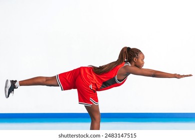 Horizontal photo a young female athlete in a red sports uniform demonstrates a balance exercise, extending one leg backward and arms forward for stability. The exercise showcases her focus, strength. - Powered by Shutterstock