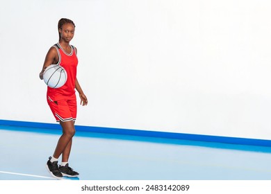 Horizontal photo a young female basketball player in a red uniform practices dribbling on an indoor court, showcasing her focus and skill in handling the ball during a training session or game. - Powered by Shutterstock