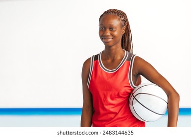 Horizontal photo a young female basketball player in a red uniform smiles confidently while holding a basketball under her arm on an indoor court, highlighting her passion and enthusiasm for the sport - Powered by Shutterstock