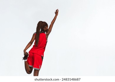 Horizontal photo a young female basketball player in a red jersey performs a standing quad stretch, reaching one arm upward. The image captures her from behind, showcasing her athleticism. - Powered by Shutterstock