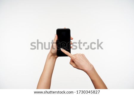 Horizontal photo of young woman's hands being raised while keeping mobile phone and swiping with forefinger on screen, posing over white background