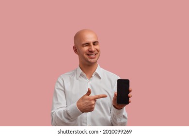 Horizontal photoshot of funny bald man with bristle in white shirt, winks and points to blank of smartphone display, recommend product. looks happy, glad to help you. Isolated on pink background