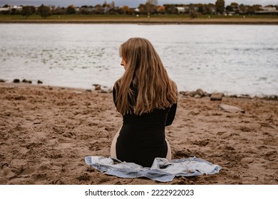 Horizontal picture of a blond woman sitting on the riverside beach; the girl is sitting on her jacket showing her back to the camera - Shutterstock ID 2222922023