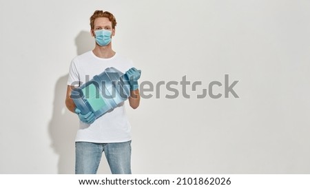 Horizontal picture of a young deliveryman holding big bottle of water, he is standing over the white background and wearing white T-shirt with blue jeans
