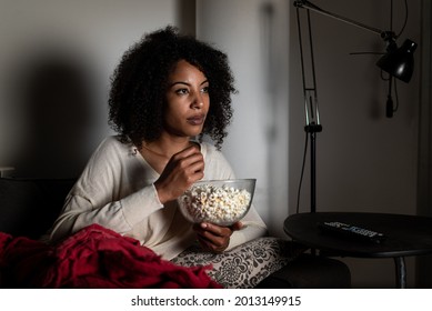 Horizontal Portrait Of An African American Woman Sitting On A Sofa. Face Is Lit Up With TV Light. She Is Eating Popcorn