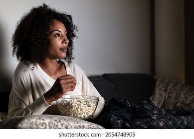 Horizontal Portrait Of An African American Woman Sitting On A Sofa. Face Is Lit Up With TV Light. She Is Holding A Bowl Of Popcorn