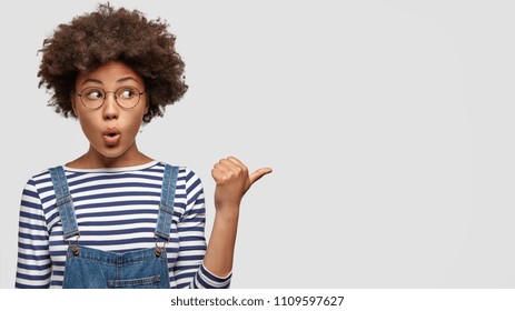 Horizontal portrait of amazed African American female youngster has puzzled and frightened expression, looks surprisingly aside, indicates with thumb at blank space. Advertisement and people.