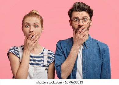 Horizontal portrait of amazed beautiful female and her boyfriend, look with astonished expressions at camera, cover mouth, can`t believe in something, isolated on pink background. Surprised friends