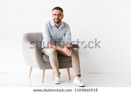 Horizontal portrait of attractive caucasian guy wearing stylish formal clothing sitting in armchair and looking at camera isolated over white background